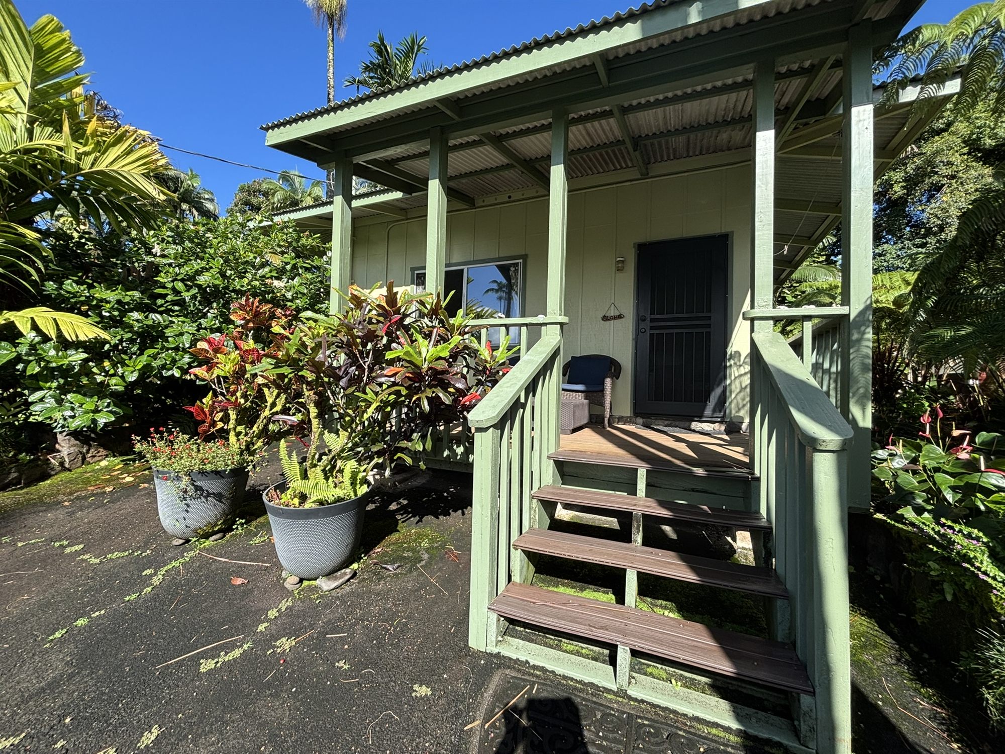The bungalow's green covered lanai and front steps in morning sun, surrounded by palms and flowering plants